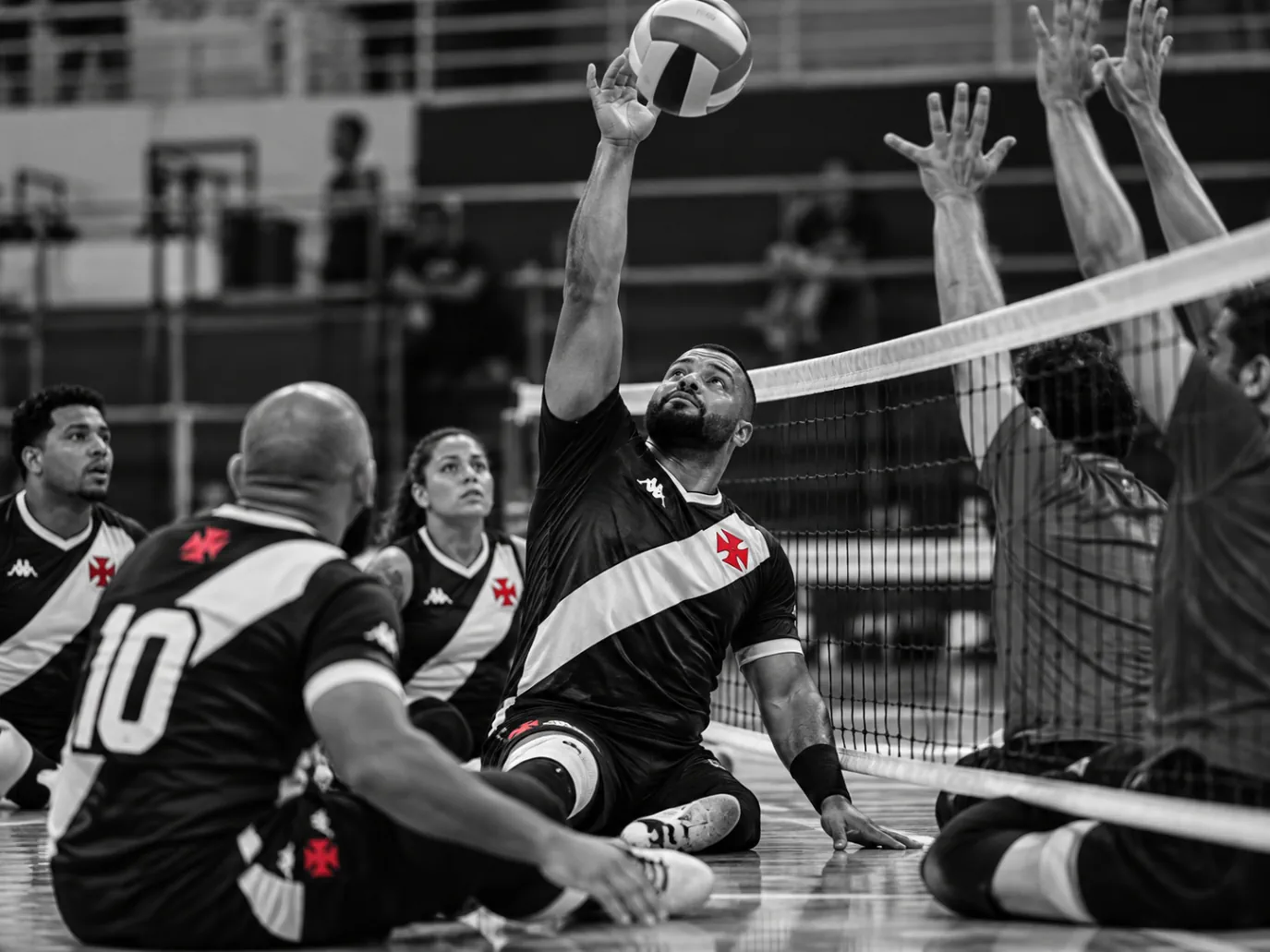 Atleta sentado na quadra atacando próximo à rede, com uniforme vascaíno